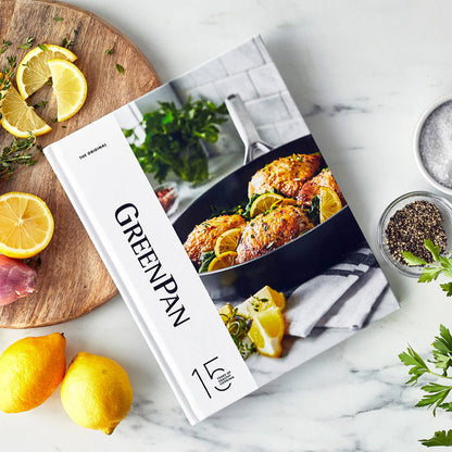 Hardcover 15-Year Anniversary Cookbook on marble with lemons, fresh herbs, salt, and pepper nearby.