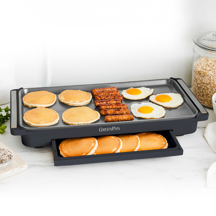 A family brunch cooks on the Advanced Griddle with Warming Drawer | Graphite—pancakes, sausage, and eggs sizzle on top while pancakes stay warm below. A jar of oats and fresh greens rest nearby.