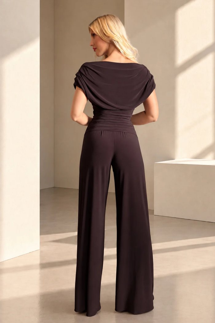 Woman viewed from behind wearing a dark brown draped wide leg jumpsuit in a sunlit room