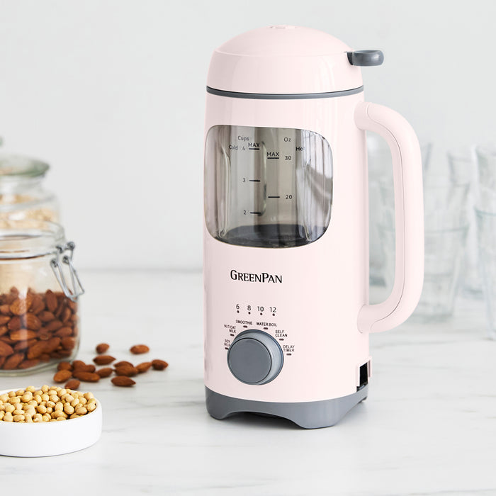 Pink milk maker with 'GreenPan' branding on a white surface with jars of nuts and seeds.