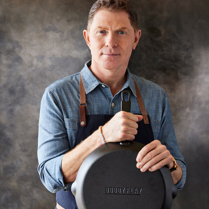 Man in blue shirt and apron holding a set of two black cast iron frypans against a gray background.