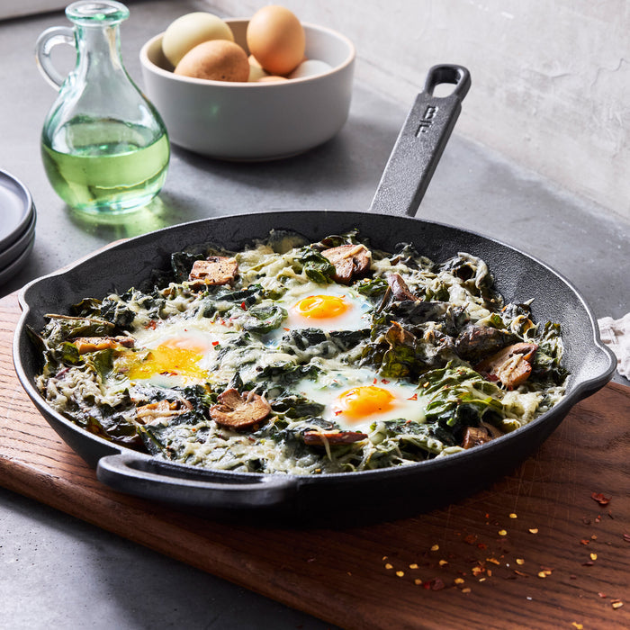 Two black cast iron frypans with baked eggs, greens, and mushrooms on a wooden board. Set includes 10 and 12 pans.