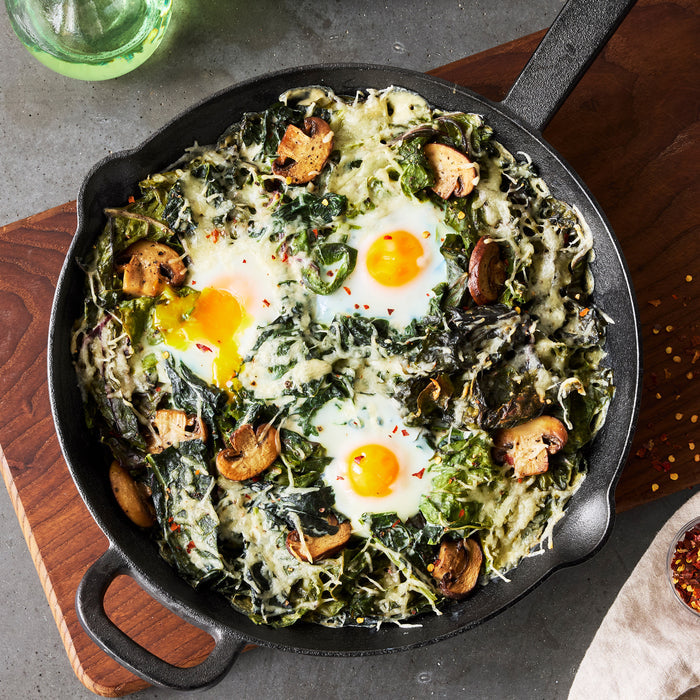 Two black cast iron frypans with baked eggs, greens, mushrooms, and cheese on a wooden board.