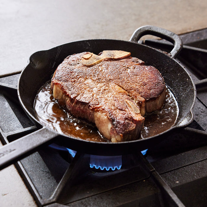 Steak cooking in a black cast iron frypan on a gas stove in a modern kitchen.