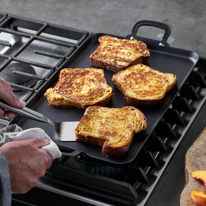 Four slices of French toast on a large black carbon steel double burner griddle, with one being flipped by a spatula.