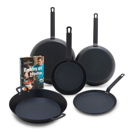 Five-piece carbon steel cookware set with Bobby at Home cookbook on a white background.