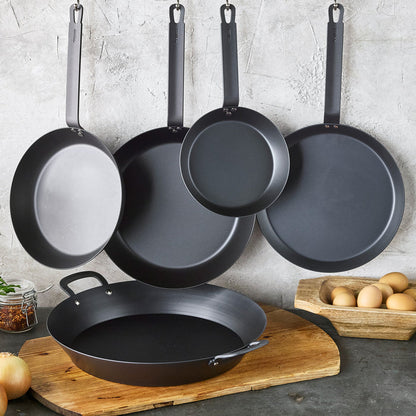 Five-piece carbon steel cookware set by GreenPan hanging on wall, cookbook visible nearby.