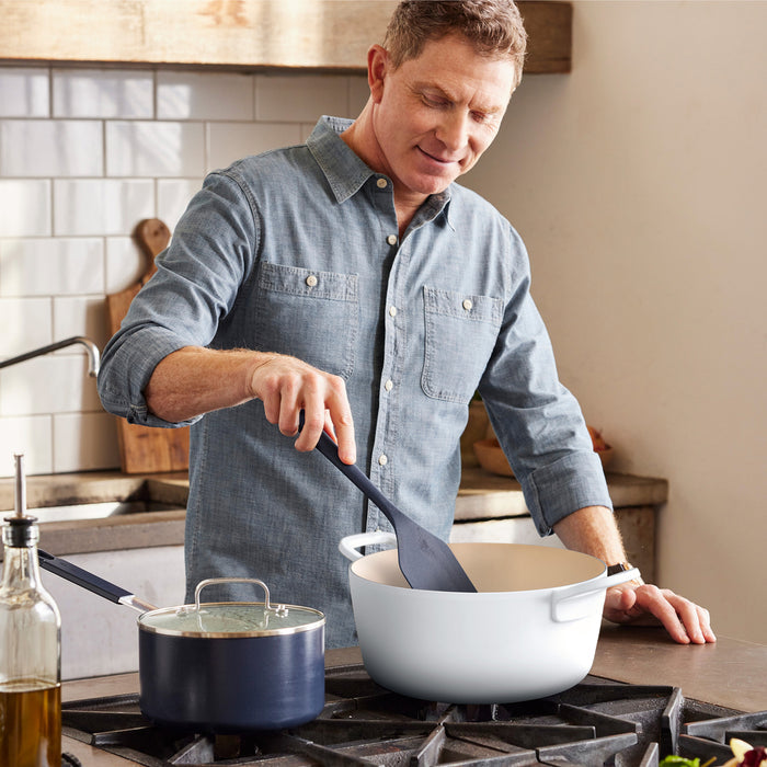 Man stirring food in a 5.5-quart ivory Dutch oven on a stove in a rustic kitchen.