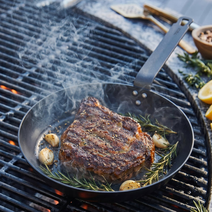 Steak with rosemary and garlic cooking in a Bobby Flay by GreenPan carbon steel frypan, smoke rising from the pan.