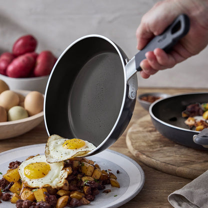 Ceramic nonstick pan with two fried eggs being served onto a plate with potatoes and meat.