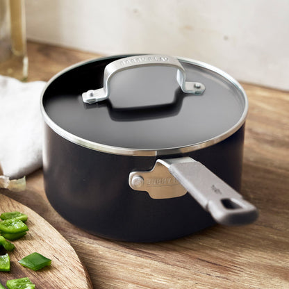Black saucepan with lid next to chopped green peppers on a wooden counter.