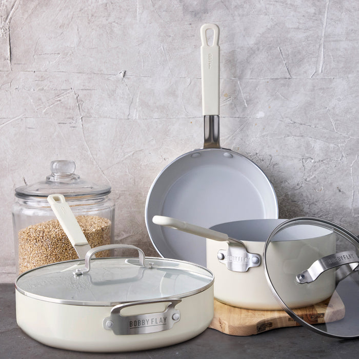 Cream ceramic nonstick cookware set with glass lids arranged on a gray countertop.