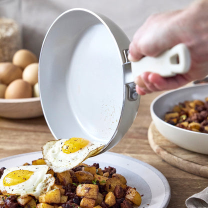 Hand slides a fried egg from a white GreenPan skillet onto potatoes and meat.