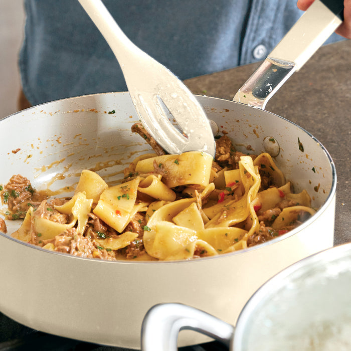 Noodles, sauce, and meat being stirred in a white ceramic nonstick pot with stainless steel handles.