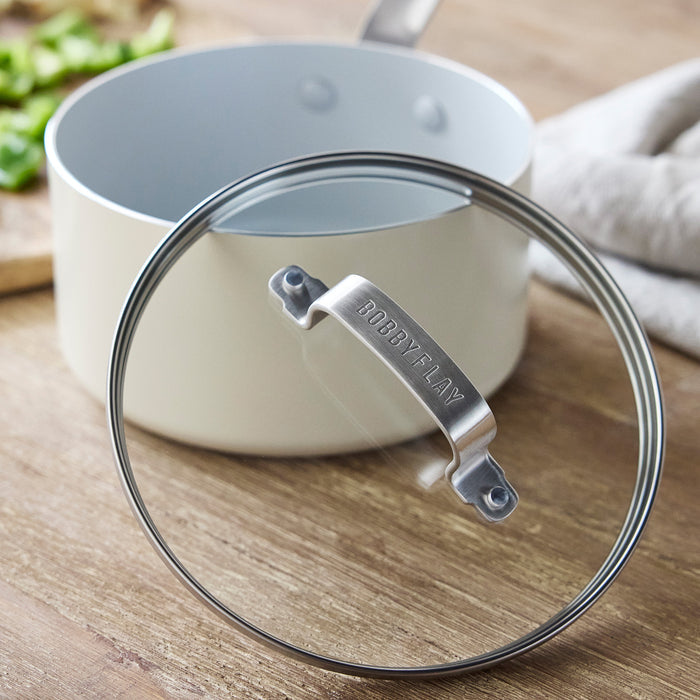 Cream ceramic saucepan with glass lid on wooden surface beside chopped green peppers.