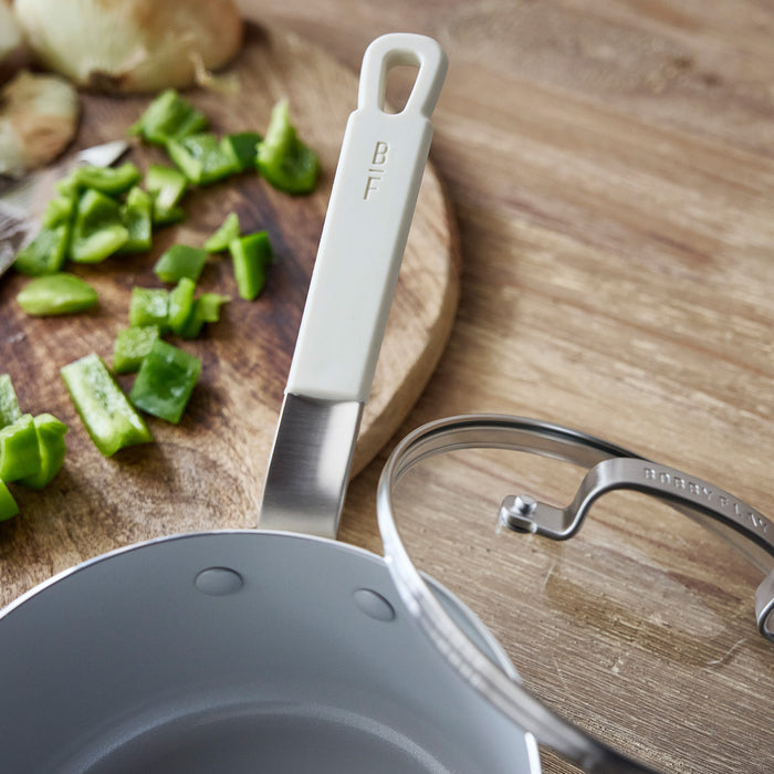 Cream ceramic nonstick cookware set with glass lid next to chopped green peppers.