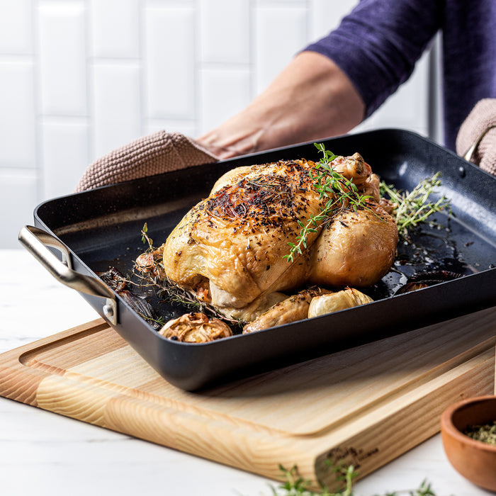 Person in oven mitts holding a 13.8″ x 10″ ceramic nonstick rectangular roasting pan containing a whole herb-roasted chicken.