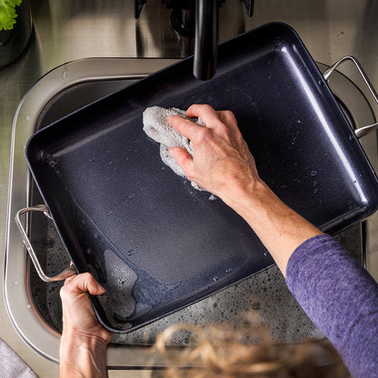 Dark ceramic nonstick 13.8″ x 10″ rectangular roasting pan being hand-washed with a sponge in a kitchen sink.