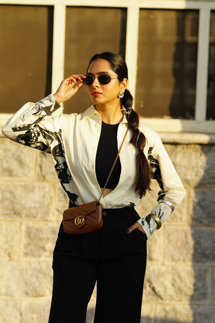 Woman wearing the CM shirt with black trousers, outdoors, hand raised to her sunglasses