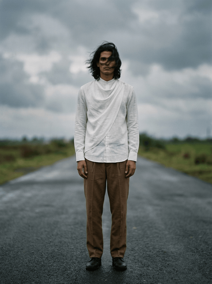 Man wearing the white Cowl shirt with brown trousers, full length front view, standing on a road under cloudy sky