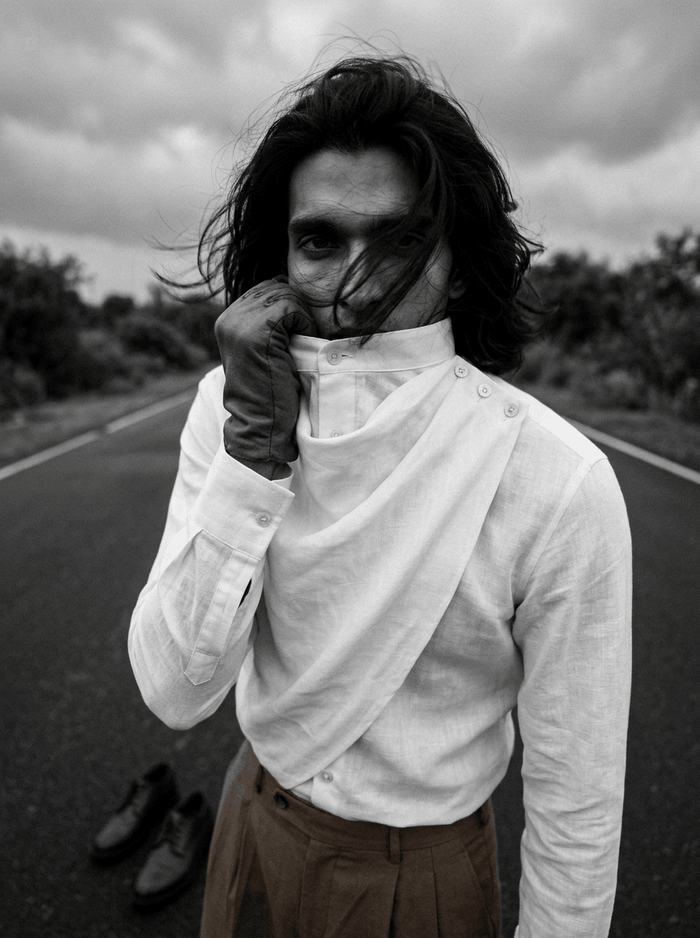 Black and white photo of a man wearing the Cowl shirt, hand at the cowl neckline, on a road
