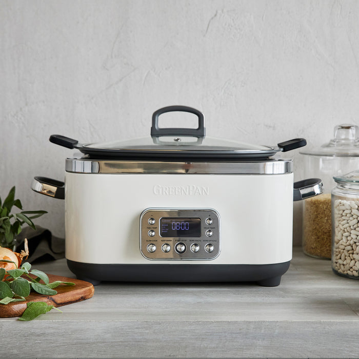 An Elite 14-in-1 6-Quart Slow Cooker in Cloud Cream with a digital display and easy presets sits on a grey countertop, surrounded by beans, onions, herbs, and a glass jar.
