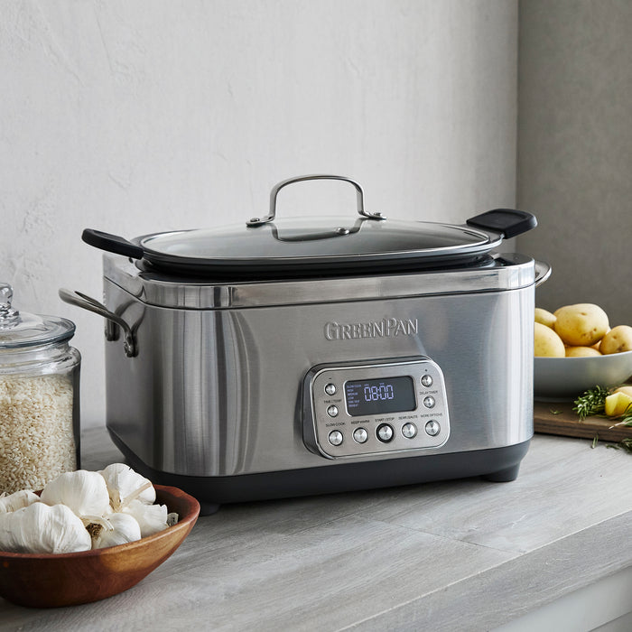 The Elite 14-in-1 6-Quart Slow Cooker | Premiere Stainless Steel with user-friendly presets sits on a kitchen counter beside a bowl of garlic, a glass jar of rice, and a plate of potatoes and herbs.