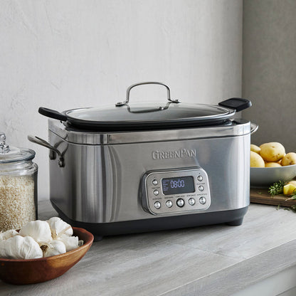 The Elite 14-in-1 6-Quart Slow Cooker | Premiere Stainless Steel with user-friendly presets sits on a kitchen counter beside a bowl of garlic, a glass jar of rice, and a plate of potatoes and herbs.