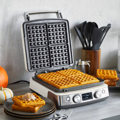 Cream-colored ceramic 4-square waffle maker on a kitchen counter, making Belgian waffles, with plates and pumpkins nearby.