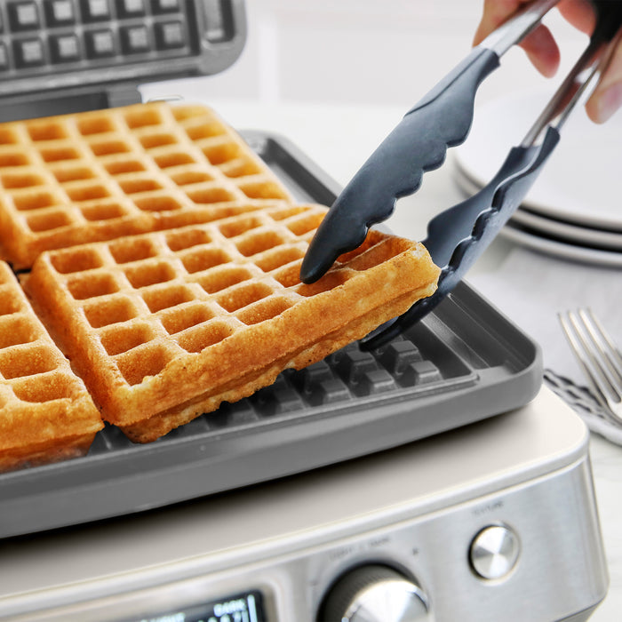 A hand lifts a golden waffle from a white ceramic 4-square waffle maker, with more waffles inside.