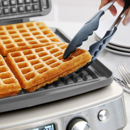 A hand lifts a golden waffle from a white ceramic 4-square waffle maker, with more waffles inside.