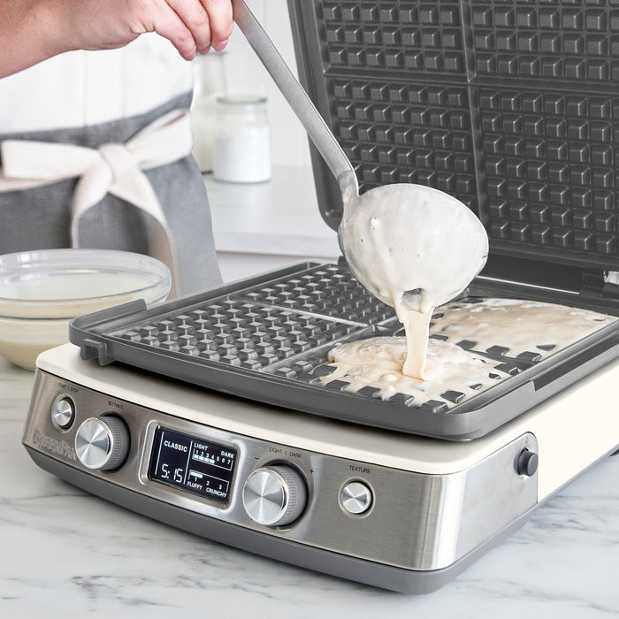 Batter being poured onto a white ceramic 4-square waffle maker on a kitchen counter.