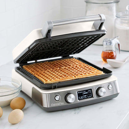 Ceramic 4-square waffle maker in cream with Liège waffles, eggs, batter, and syrup on the counter.