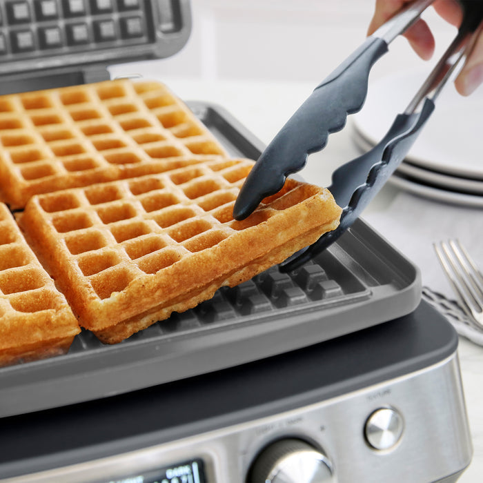 Waffle maker in graphite with hand using tongs to remove waffles; four waffles visible on nonstick ceramic plates.