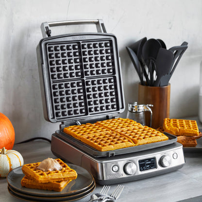 4-square ceramic waffle maker on a counter with pumpkin waffles, utensils, and small pumpkins nearby.