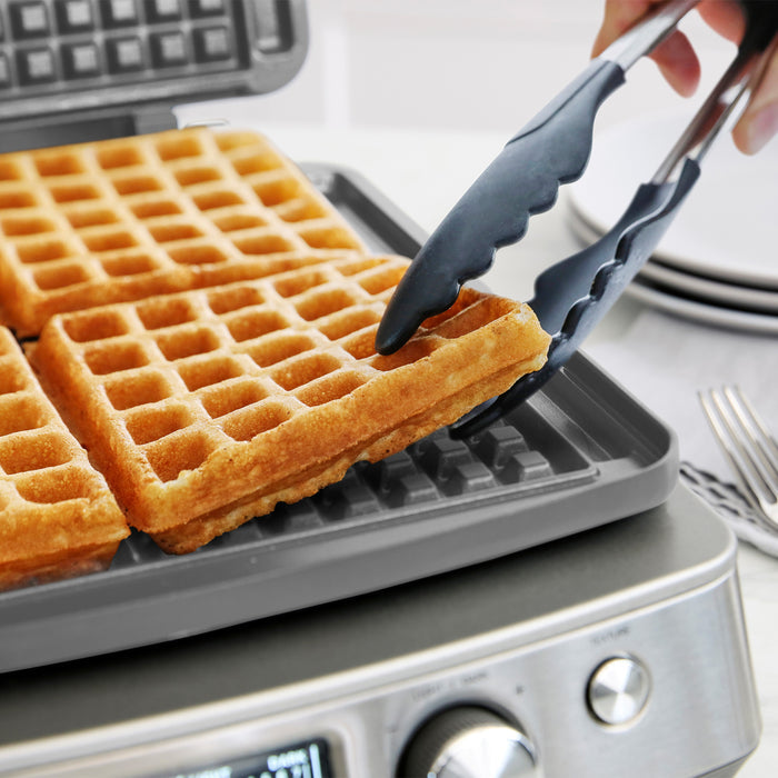 Using tongs, someone lifts a golden Belgian waffle from the Elite Ceramic Nonstick 4-Square Waffle Maker | Premiere Stainless Steel, with fresh waffles inside and plates and utensils visible in the background.