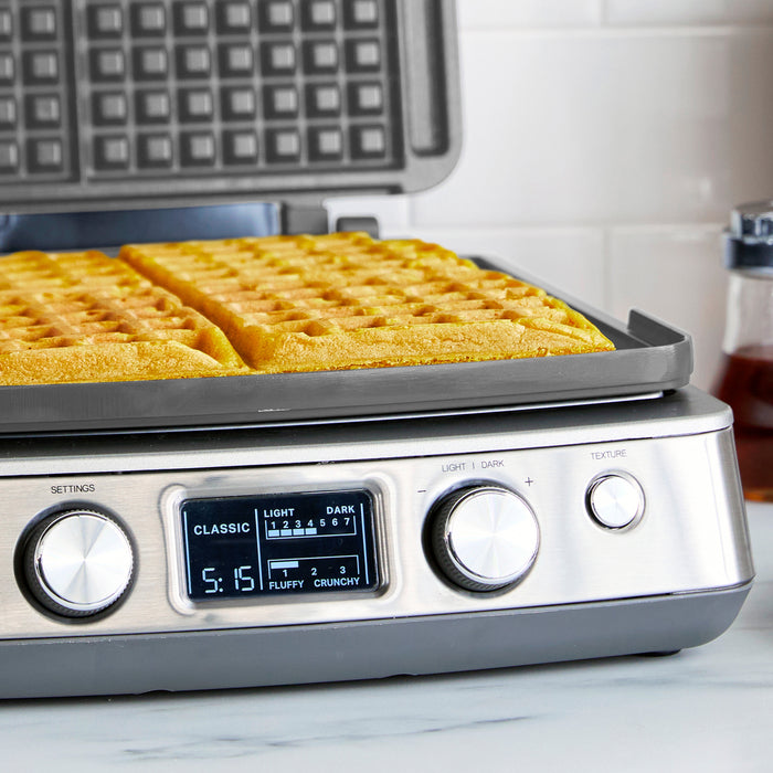 The Elite Ceramic Nonstick 4-Square Waffle Maker | Premiere Stainless Steel sits on a countertop with two golden Belgian waffles inside. Its digital display offers settings for light, dark, classic, texture, plus dials and buttons below.