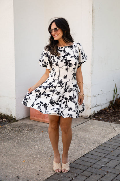 Woman wearing a black and white floral dress standing against a white wall.