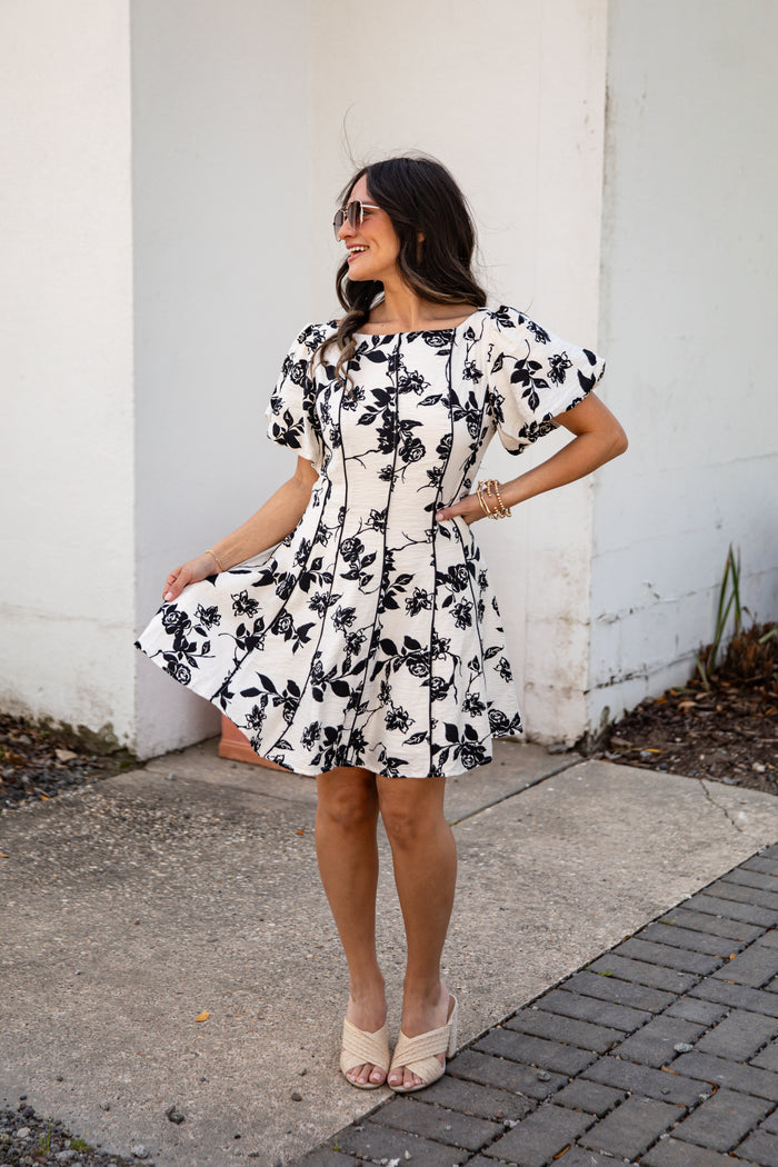 Woman wearing a black and white floral dress standing against a white wall.