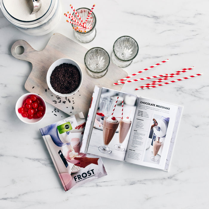Smoothies, milkshake ingredients, glasses, and straws arranged beside a Frost Classic recipe book on marble surface.
