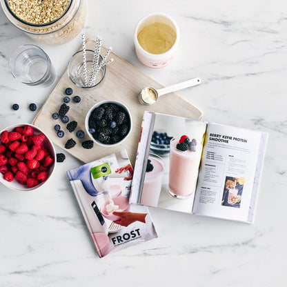 Fresh berries, a scoop of protein powder, and the Frost Classic Recipe Book on a white marble countertop.