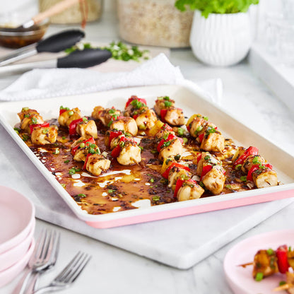 Sheet pan of skewered chicken and vegetables on a white surface with utensils and plates.