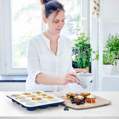 Woman icing muffins beside a navy GreenLife ceramic muffin pan on a kitchen counter with houseplants.
