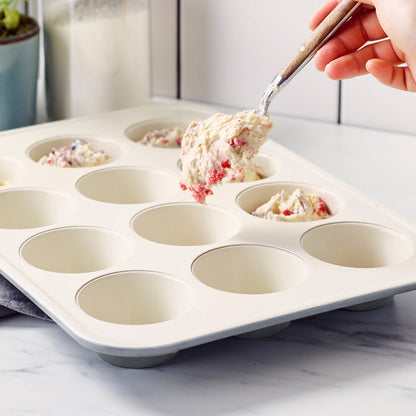 Hand scooping muffin batter into a gray ceramic nonstick muffin pan on a kitchen counter.