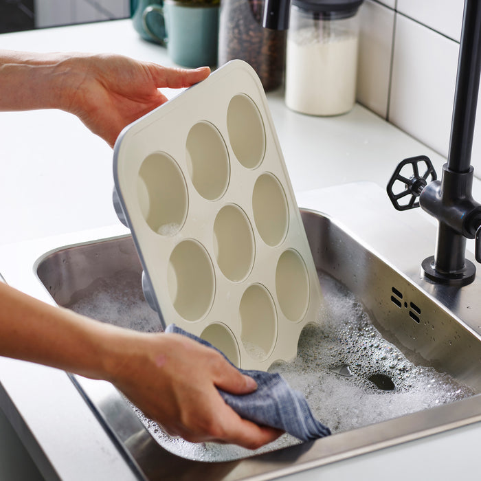 Washing a gray ceramic muffin pan with a cloth in a soapy sink.