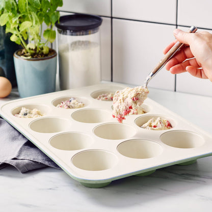 Sage green ceramic muffin pan with batter being scooped in, shown on a kitchen counter.