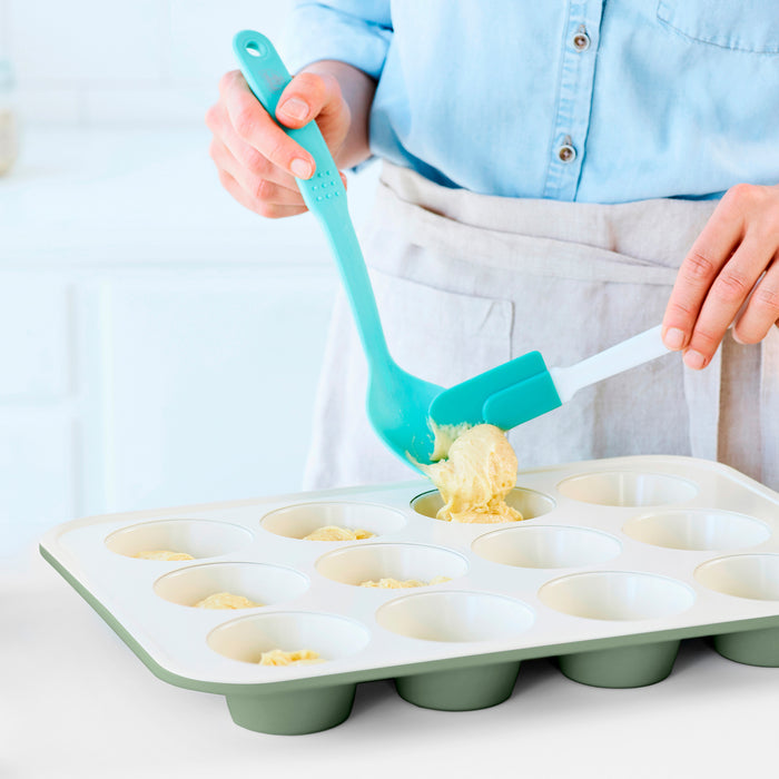 Batter being scooped into a sage green ceramic nonstick muffin pan in a bright kitchen.