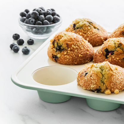 Sage green ceramic muffin pan with six blueberry muffins and a bowl of blueberries on a white surface.