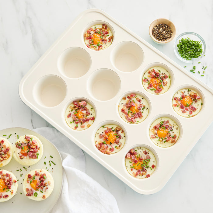 Sage green ceramic nonstick muffin pan with egg bites topped with bacon, herbs, and cheese on a white surface.