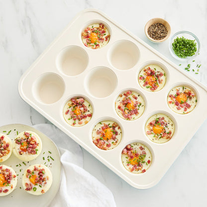 Sage green ceramic nonstick muffin pan with egg bites topped with bacon, herbs, and cheese on a white surface.